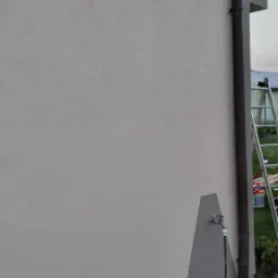 Fragment ściany budynku z nową, jasną elewacją i fragmentem pomalowanym na szaro, widoczna rynna, drabina i kran z wężem ogrodowym.