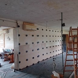 Usługi remontowo-wykończeniowe - Wnętrze w trakcie prac: ściana z płyt OSB z widocznymi kołkami mocującymi, zabezpieczona folią podłoga, drabina i narzędzia. W tle widoczny klimatyzator i pomieszczenie z wyposażeniem.