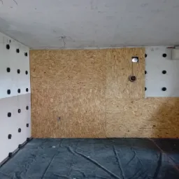 Wnętrze w trakcie ocieplania poddasza: ściany z przymocowaną izolacją z czarnymi kołkami, fragment ściany wykończony płytą OSB z otworem, podłoga zabezpieczona folią.