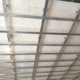 Usługi remontowo-wykończeniowe - Metalowa konstrukcja sufitu podwieszanego z widocznymi elementami mocującymi, surowy tynk na stropie, wisząca żarówka.