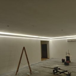 Usługi remontowo-wykończeniowe - Wnętrze pokoju w trakcie prac wykończeniowych, zabezpieczona folią podłoga, drabina drewniana, narzędzia malarskie, świeżo pomalowane ściany z zamontowanym oświetleniem LED pod sufitem i klimatyzacją.