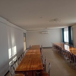 Usługi remontowo-wykończeniowe - Wnętrze sali w Mrozach po remoncie: Długie drewniane stoły z krzesłami ustawione w rzędach, białe ściany z promieniami słońca, klimatyzacja i dekoracyjne oświetlenie sufitowe.