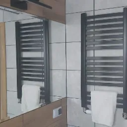 Nowoczesna łazienka z umywalką nablatową, grzejnikiem drabinkowym w kolorze antracytowym z powieszonym białym ręcznikiem na tle szarych, kwadratowych płytek i drewnopodobnej zabudowy meblowej.