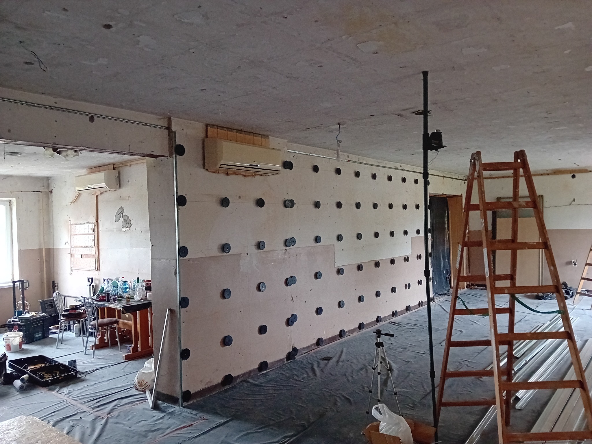 Wnętrze w trakcie prac: ściana z płyt OSB z widocznymi kołkami mocującymi, zabezpieczona folią podłoga, drabina i narzędzia. W tle widoczny klimatyzator i pomieszczenie z wyposażeniem.