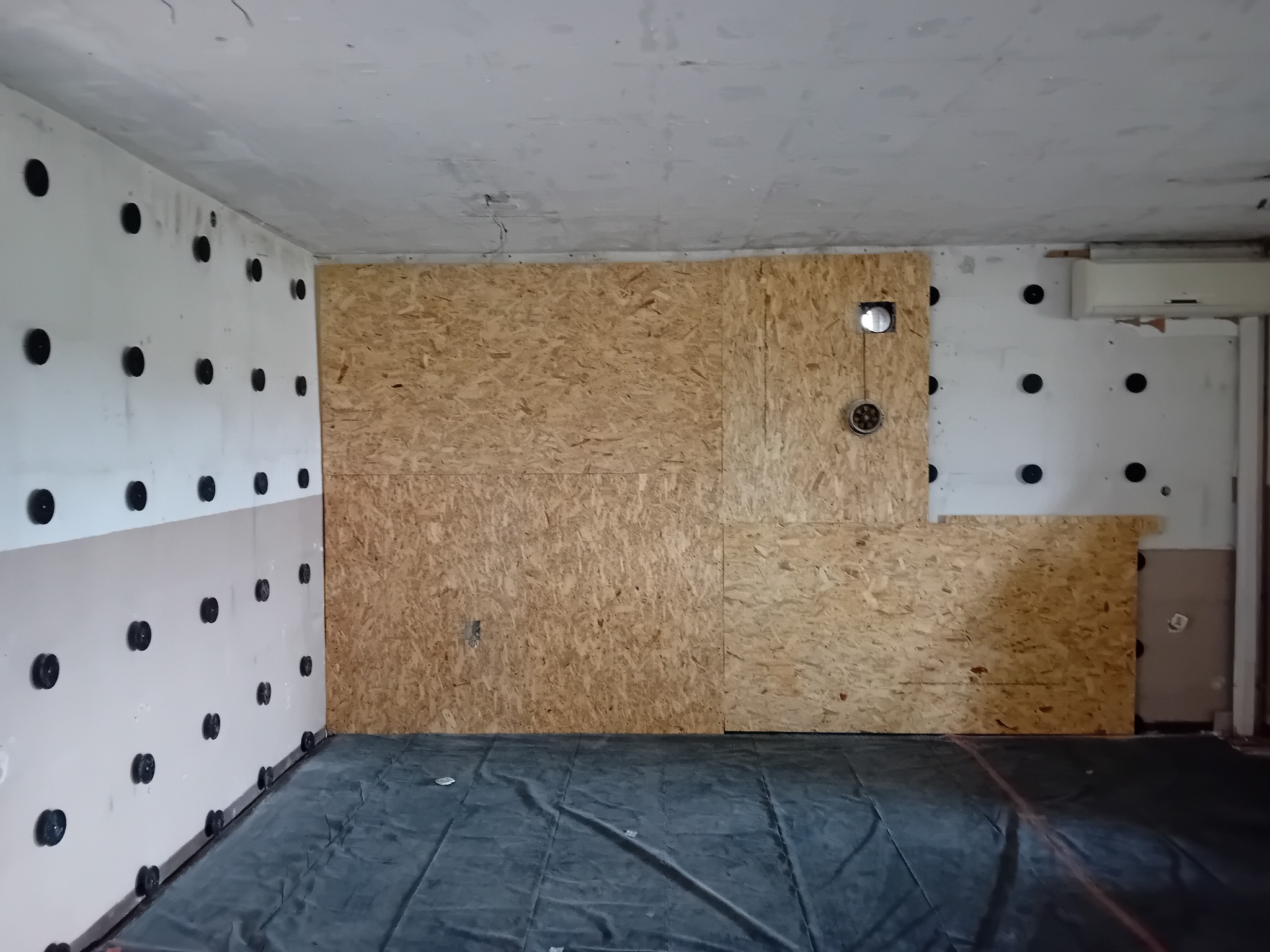 Wnętrze w trakcie ocieplania poddasza: ściany z przymocowaną izolacją z czarnymi kołkami, fragment ściany wykończony płytą OSB z otworem, podłoga zabezpieczona folią.