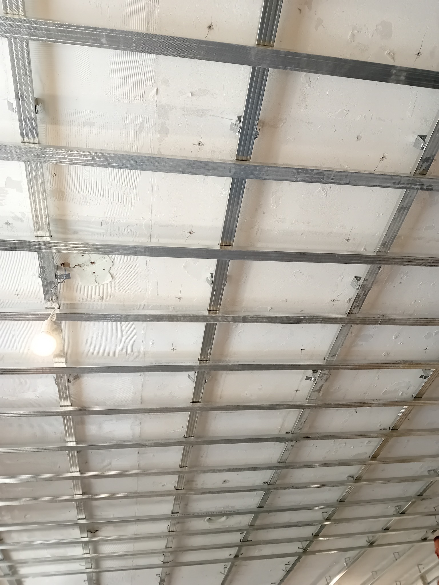 Metalowa konstrukcja sufitu podwieszanego z widocznymi elementami mocującymi, surowy tynk na stropie, wisząca żarówka.