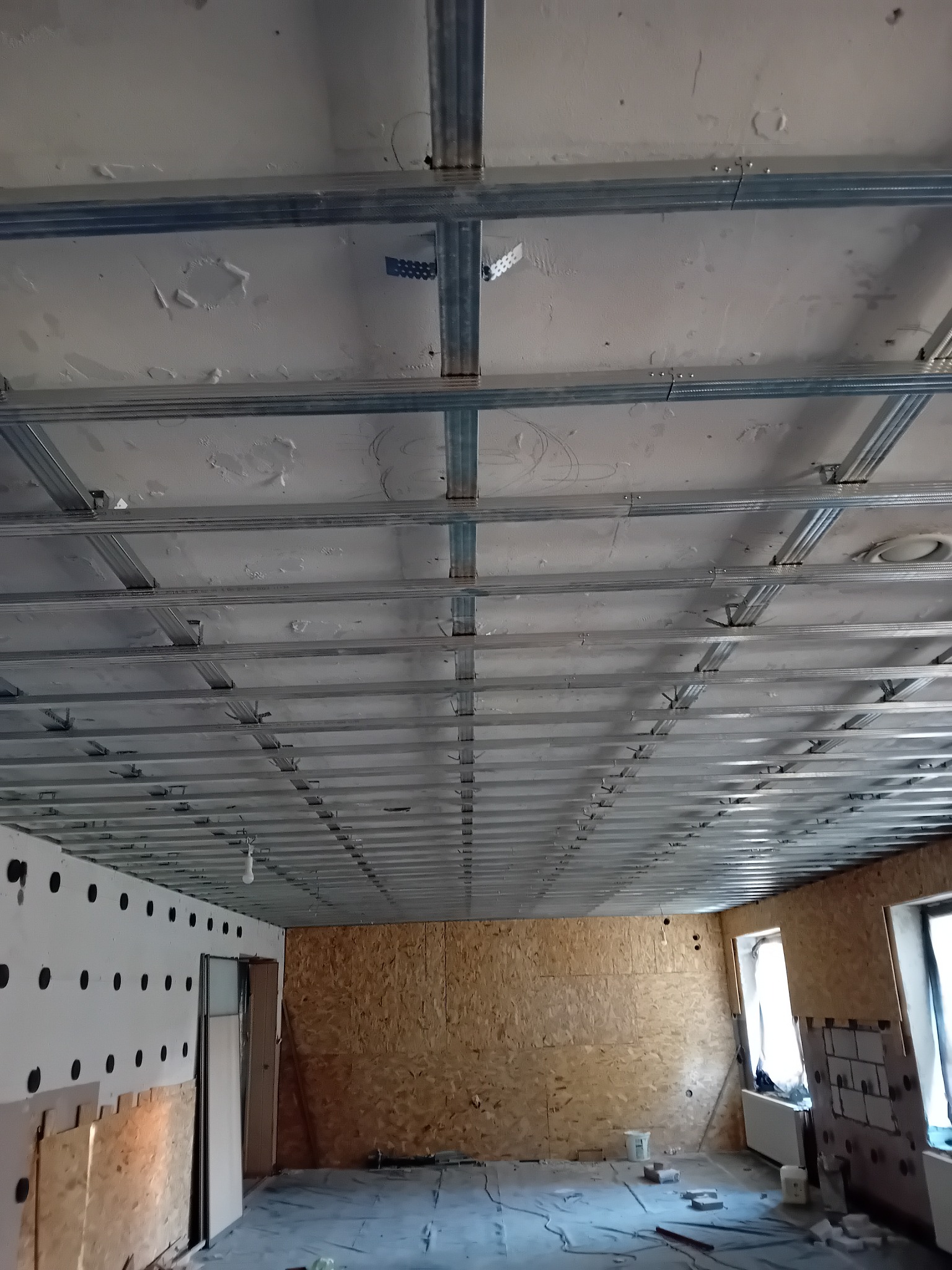 Sufit podwieszany z metalową konstrukcją w trakcie montażu, widoczne płyty OSB na ścianach i przygotowane otwory w ścianie z białej pianki.