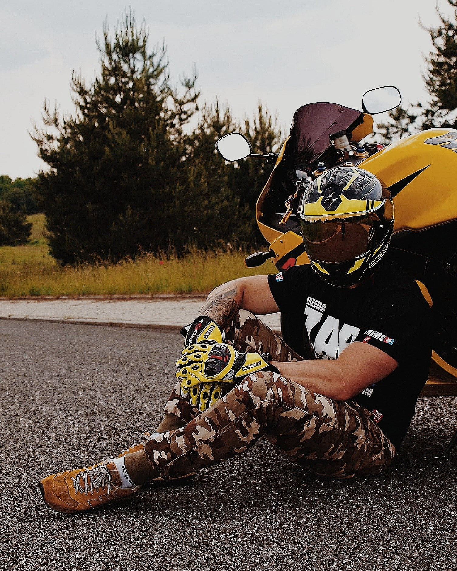 Mężczyzna w kasku i rękawicach motocyklowych siedzi na asfalcie obok żółtego motocykla sportowego, ubrany w kamuflażowe spodnie i czarną koszulkę z nadrukiem.