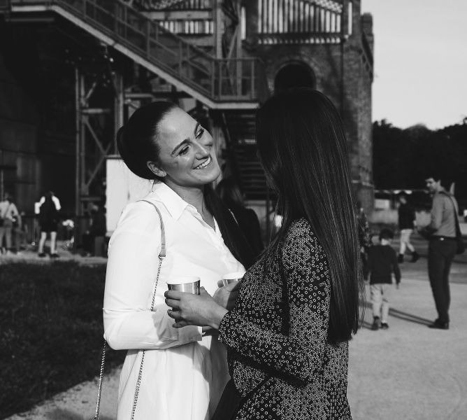 Dwie uśmiechnięte kobiety rozmawiają, trzymając kubki, na tle industrialnego budynku z metalowymi schodami i ceglaną wieżą, zdjęcie czarno-białe.