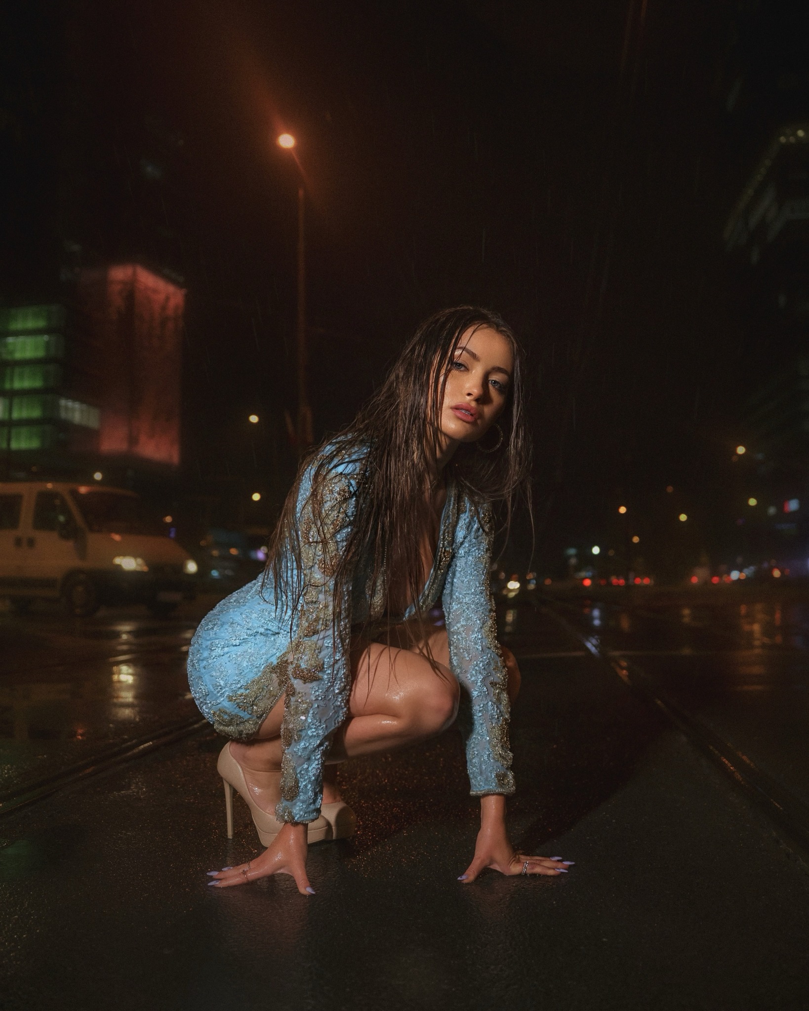Kobieta w błękitnej, zdobionej sukience i szpilkach kuca na mokrej ulicy w nocnym, deszczowym krajobrazie miejskim, z widocznymi budynkami i światłami w tle.