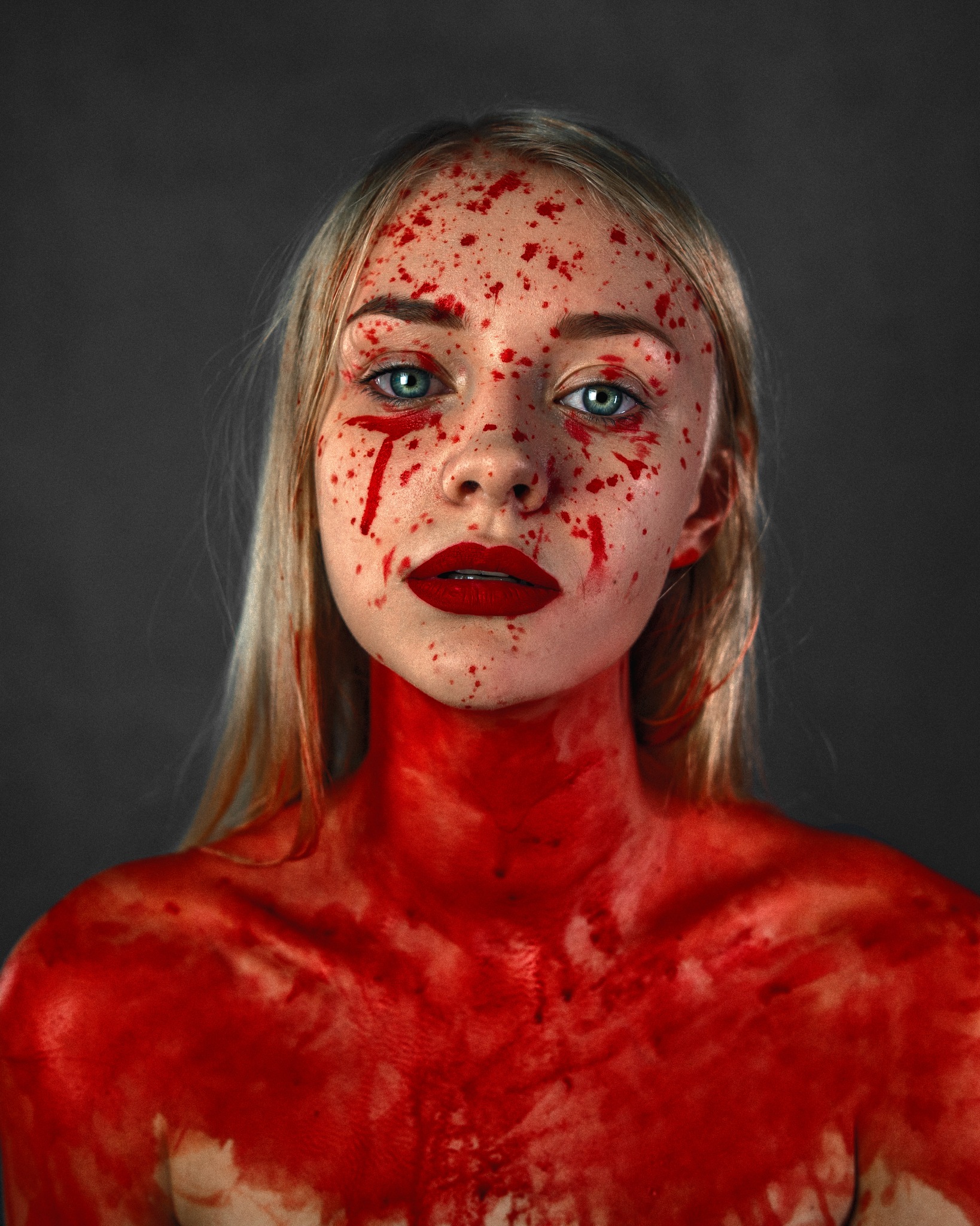 Portret kobiety z blond włosami i intensywnym makijażem ust w kolorze czerwieni, której twarz i dekolt pokryte są czerwonymi plamami przypominającymi krew, na ciemnoszarym tle.