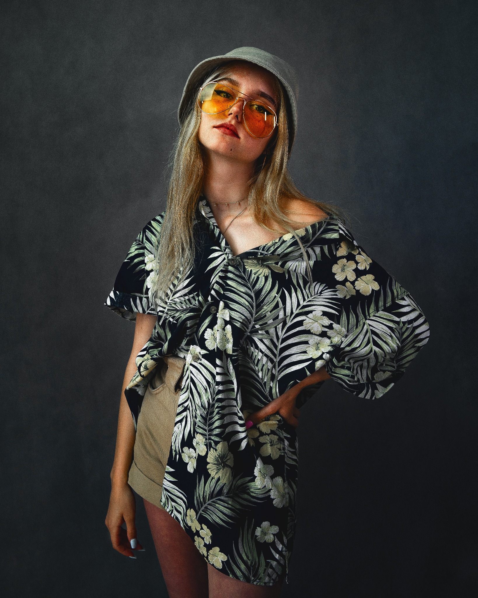 Portret młodej kobiety w kapeluszu typu bucket, dużych, żółtych okularach i kwiecistej koszuli, pozującej na ciemnym tle.