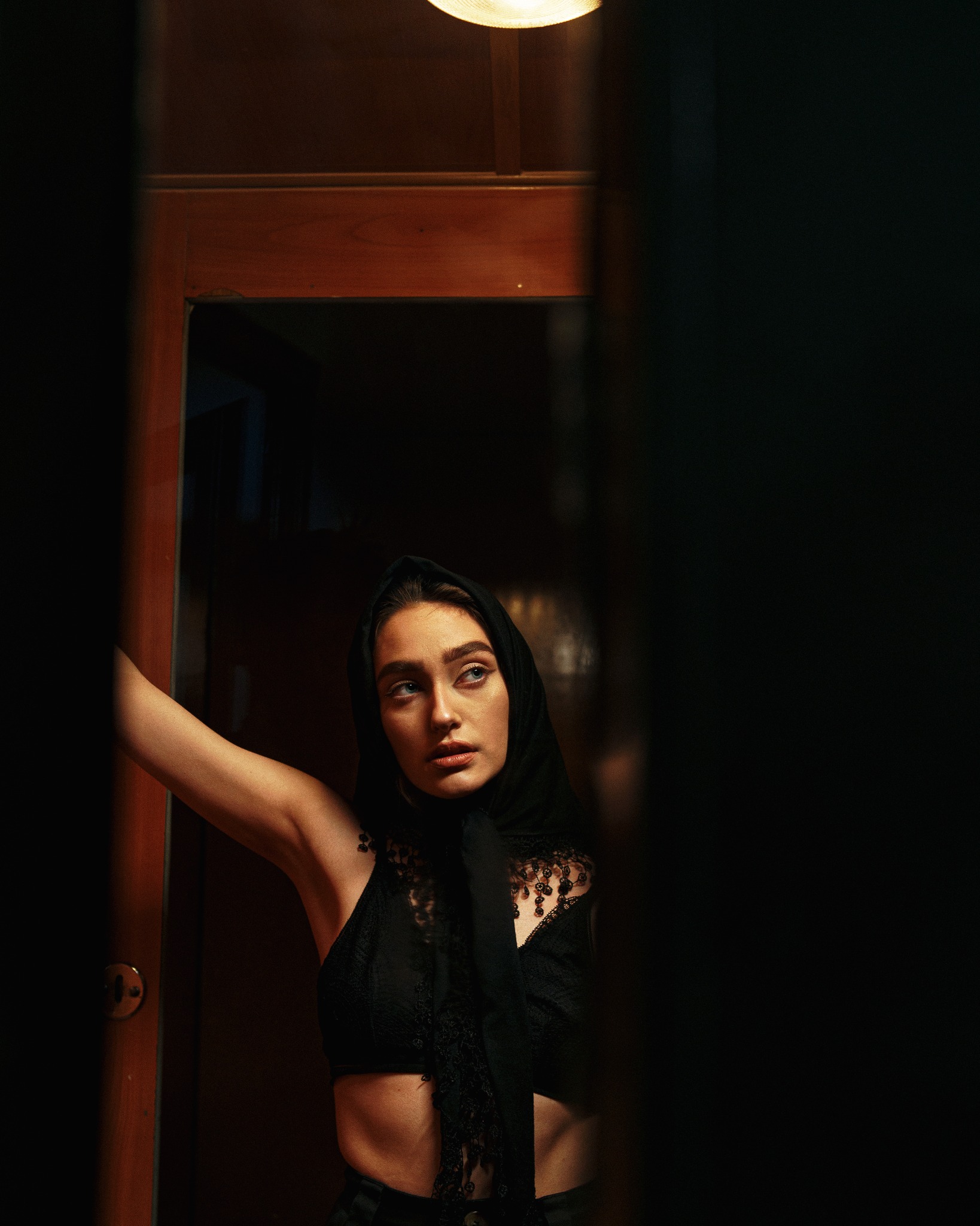 Portret kobiety w czarnej bieliźnie i chuście na głowie, stojącej w progu ciemnego pokoju, oświetlonej górnym światłem, z wyrazem zamyślenia na twarzy.