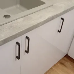 Szare blaty kuchenne imitujące beton, zintegrowany zlewozmywak, białe fronty szafek z ciemnymi, prostokątnymi uchwytami