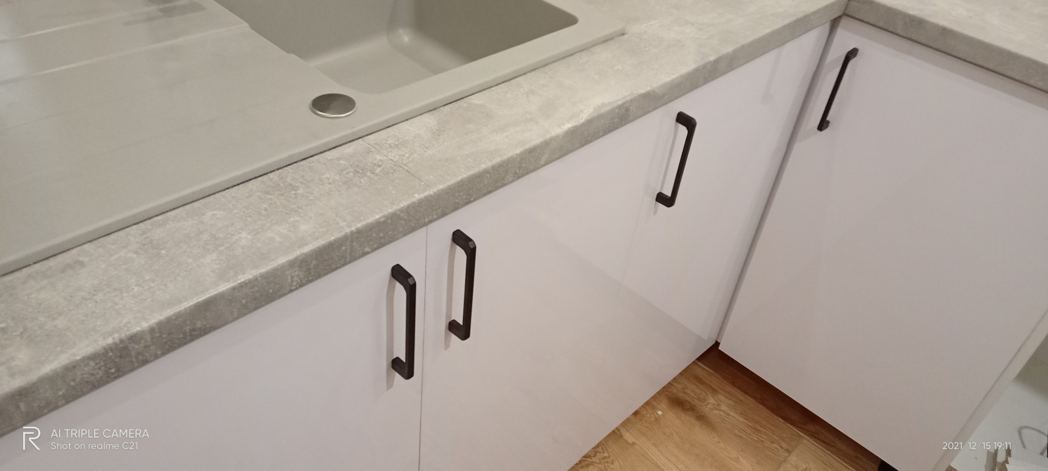 Szare blaty kuchenne imitujące beton, zintegrowany zlewozmywak, białe fronty szafek z ciemnymi, prostokątnymi uchwytami