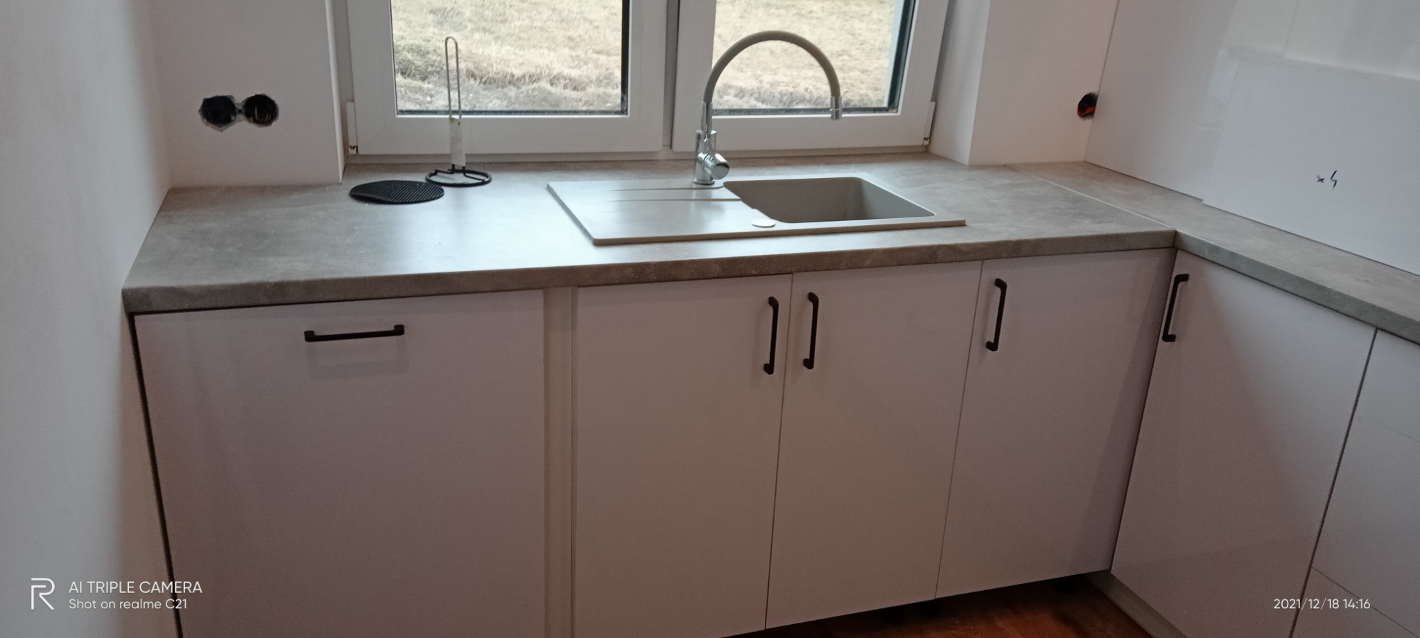 Nowoczesna kuchnia z białymi szafkami z czarnymi uchwytami, szarym blatem i zlewozmywakiem pod oknem, widoczne niedokończone gniazdka elektryczne.