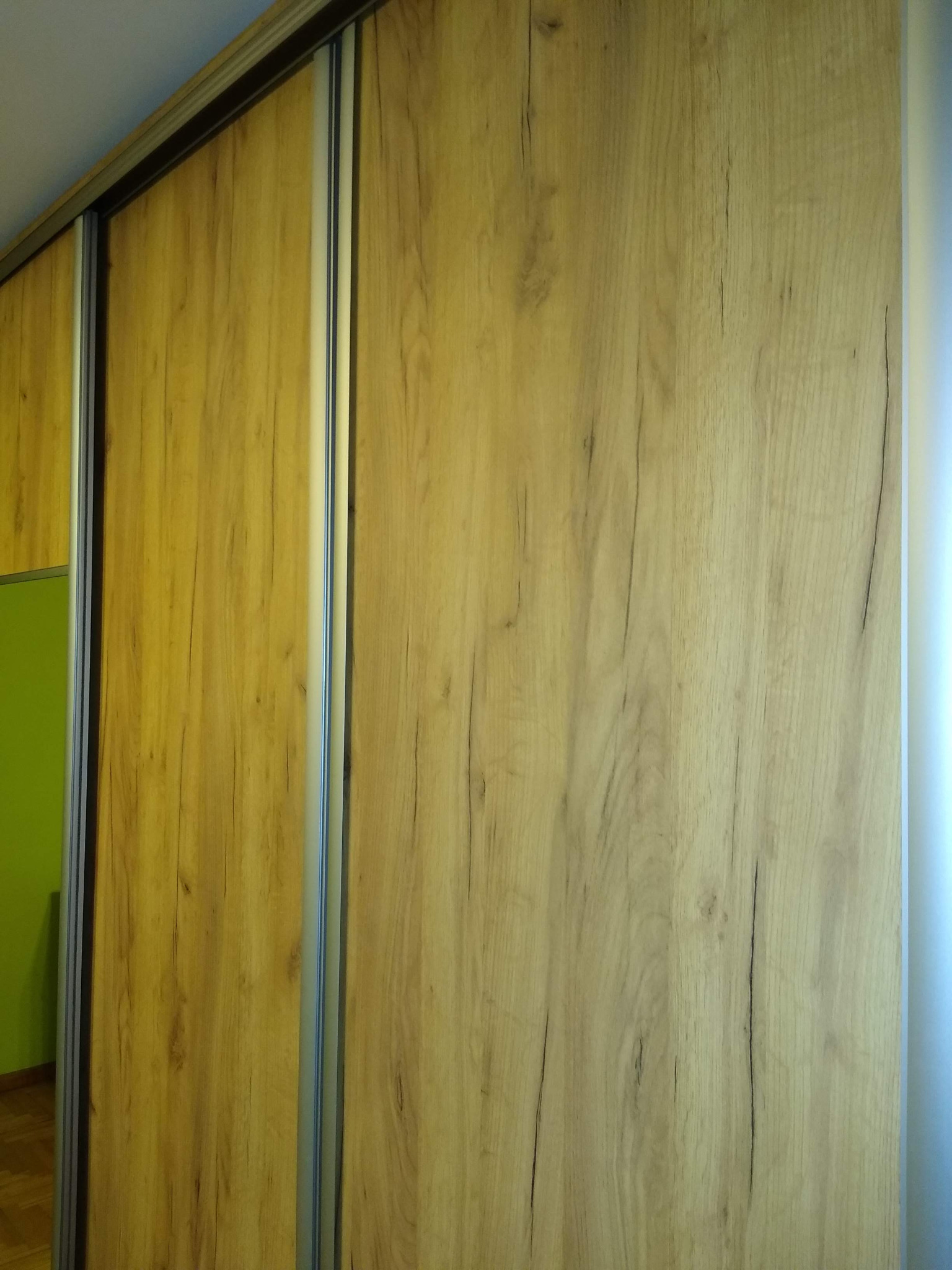Fragment szafy przesuwnej z okleiną imitującą jasne drewno, widoczny metalowy profil prowadnicy i fragment zielonej ściany wewnątrz szafy.