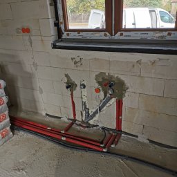 Grzegorz Latała - Świeżo wykonane podejścia instalacyjne wodno-kanalizacyjne w ścianie z betonu komórkowego, widoczne rury z tworzywa sztucznego w kolorze czerwonym i szarym, częściowo zamurowane, z widocznymi...