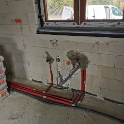 Świeżo wykonane podejścia instalacyjne wodno-kanalizacyjne w ścianie z betonu komórkowego, widoczne rury z tworzywa sztucznego w kolorze czerwonym i szarym, częściowo zamurowane, z widocznymi...