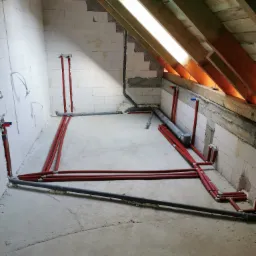 Instalacja rur grzewczych w surowym pomieszczeniu na poddaszu, widoczne czerwone rury rozprowadzone po betonowej podłodze i wyprowadzone na ściany z białych bloczków.