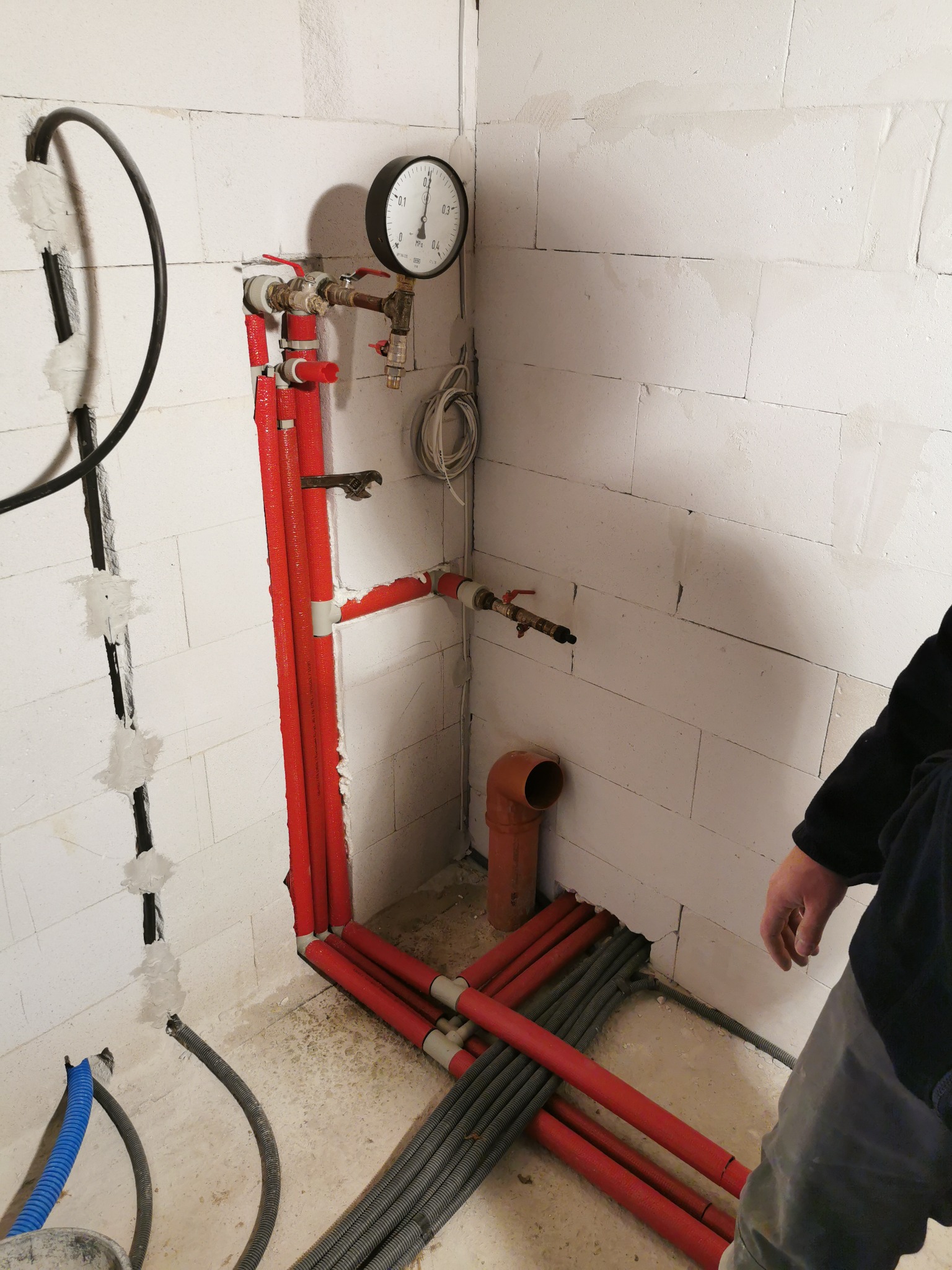 Instalacja grzewcza w trakcie montażu, widoczne czerwone rury, szare peszle, manometr przytwierdzony do ściany z betonu komórkowego, oraz fragment osoby nadzorującej prace.