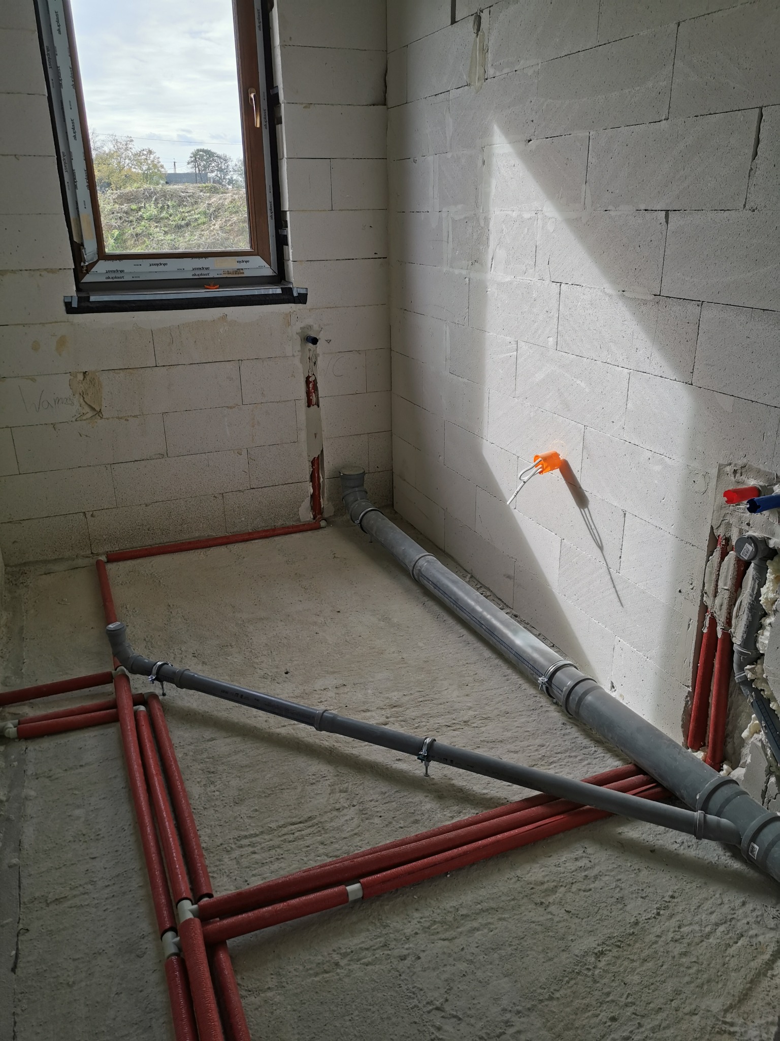 Surowy stan: Instalacja wodno-kanalizacyjna w trakcie montażu w pomieszczeniu z oknem, widoczne rury kanalizacyjne szare i rury grzewcze czerwone, ściany z białego betonu komórkowego.