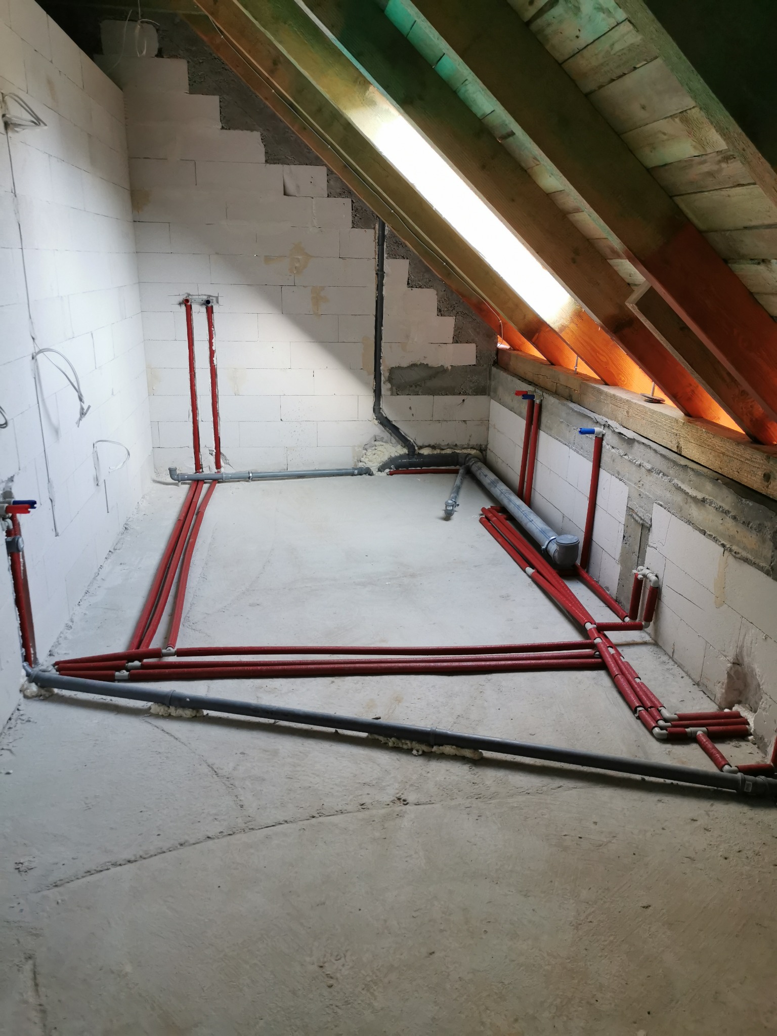Instalacja rur grzewczych w surowym pomieszczeniu na poddaszu, widoczne czerwone rury rozprowadzone po betonowej podłodze i wyprowadzone na ściany z białych bloczków.