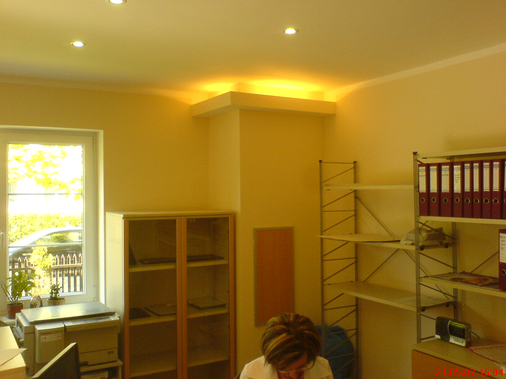 Żółte ściany biura z oknem, szafkami i półkami, oświetlenie sufitowe wnękowe.