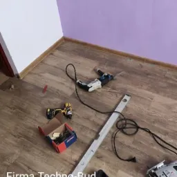 Podłoga z paneli, przy ścianie w kolorze fioletowym i białym, z narzędziami budowlanymi: wiertarka, wkrętarka, poziomica, skrzynka z wkrętami oraz wyrzynarka.