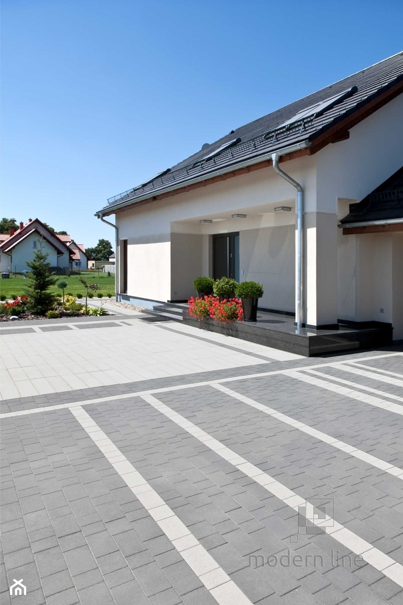 Nowoczesny dom jednorodzinny z szarą dachówką, białą elewacją i eleganckim podjazdem z kostki brukowej w odcieniach szarości i bieli, uzupełniony zielenią i czerwonymi kwiatami w donicach.