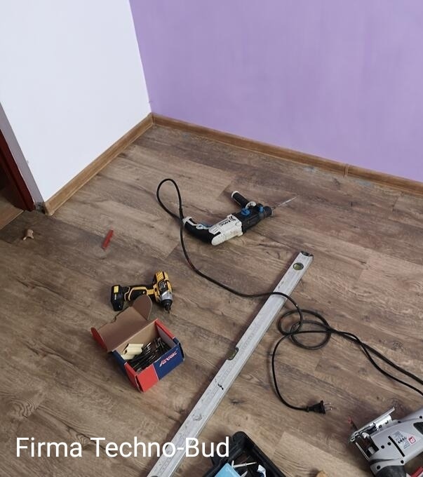 Podłoga z paneli, przy ścianie w kolorze fioletowym i białym, z narzędziami budowlanymi: wiertarka, wkrętarka, poziomica, skrzynka z wkrętami oraz wyrzynarka.