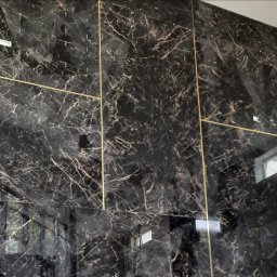 HaGieHubertGaweł - Ściana wykończona czarnymi płytami imitującymi marmur z wyraźnym, brązowym żyłowaniem i złotymi listwami, tworzącymi geometryczny wzór.