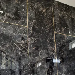 Ściana wykończona czarnymi płytami imitującymi marmur z wyraźnym, brązowym żyłowaniem i złotymi listwami, tworzącymi geometryczny wzór.
