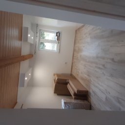 HaGieHubertGaweł - Pokój w trakcie wykańczania, z panelami na ścianie imitującymi drewno, widoczne paczki paneli na podłodze, drewniana belka stropowa i oświetlenie punktowe w suficie.