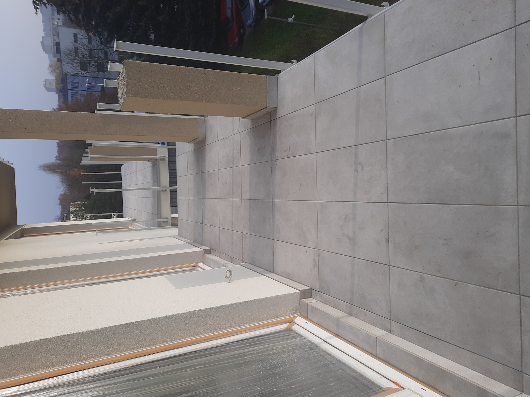 Wykończony balkon z szarymi płytkami ceramicznymi ułożonymi w regularny wzór, widoczne elementy konstrukcyjne i balustrada, w tle budynki mieszkalne.