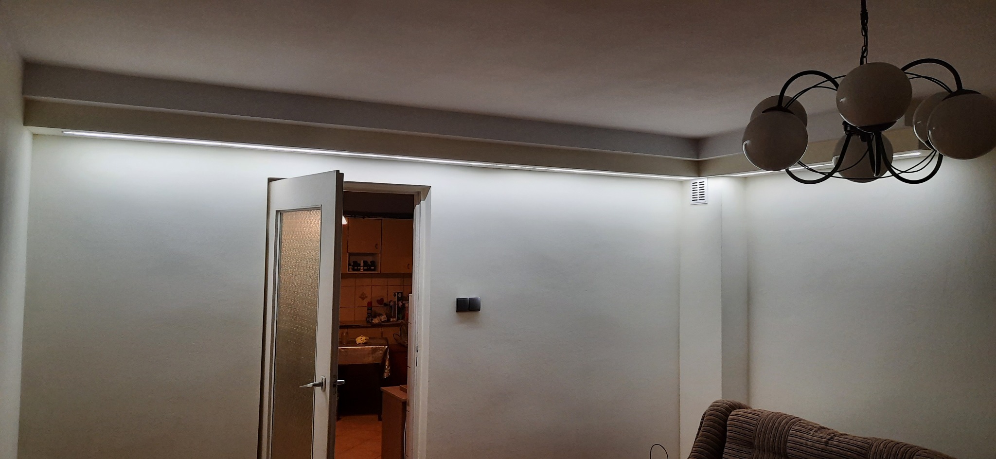 Jasne wnętrze z oświetleniem LED ukrytym w listwach sufitowych, widoczny fragment lampy wiszącej i otwarte drzwi do kuchni z widocznym wyposażeniem.