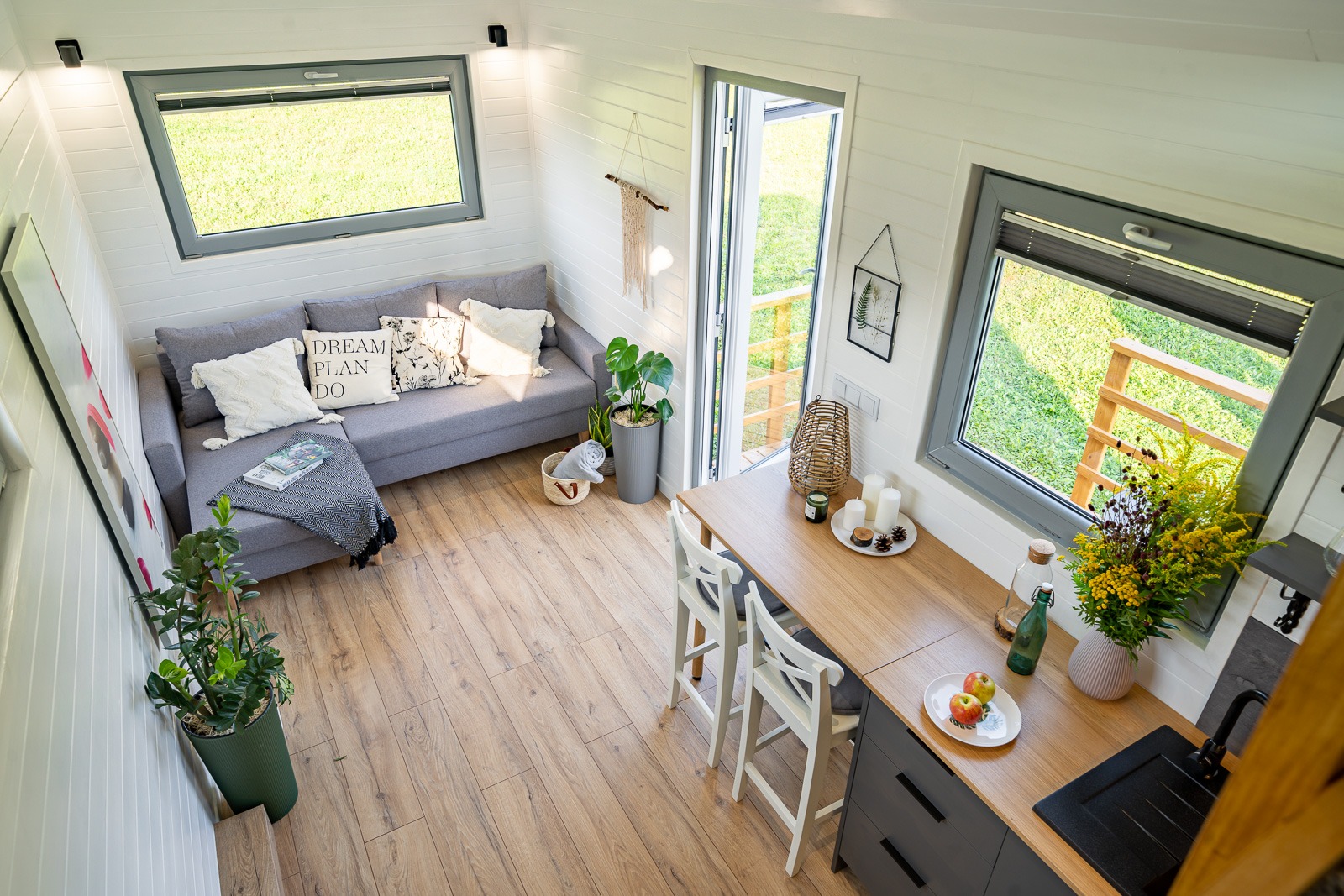 Wnętrze domu szkieletowego z widokiem z góry: szara sofa z poduszkami, drewniana podłoga, stół z białymi krzesłami i dekoracjami, widoczne okna z zielonym krajobrazem na zewnątrz.