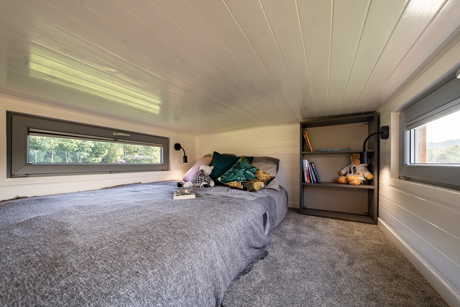 Przytulna sypialnia na poddaszu z oknami, szarą pościelą, pluszowym misiem i książkami na półce, oświetlona lampką nocną.