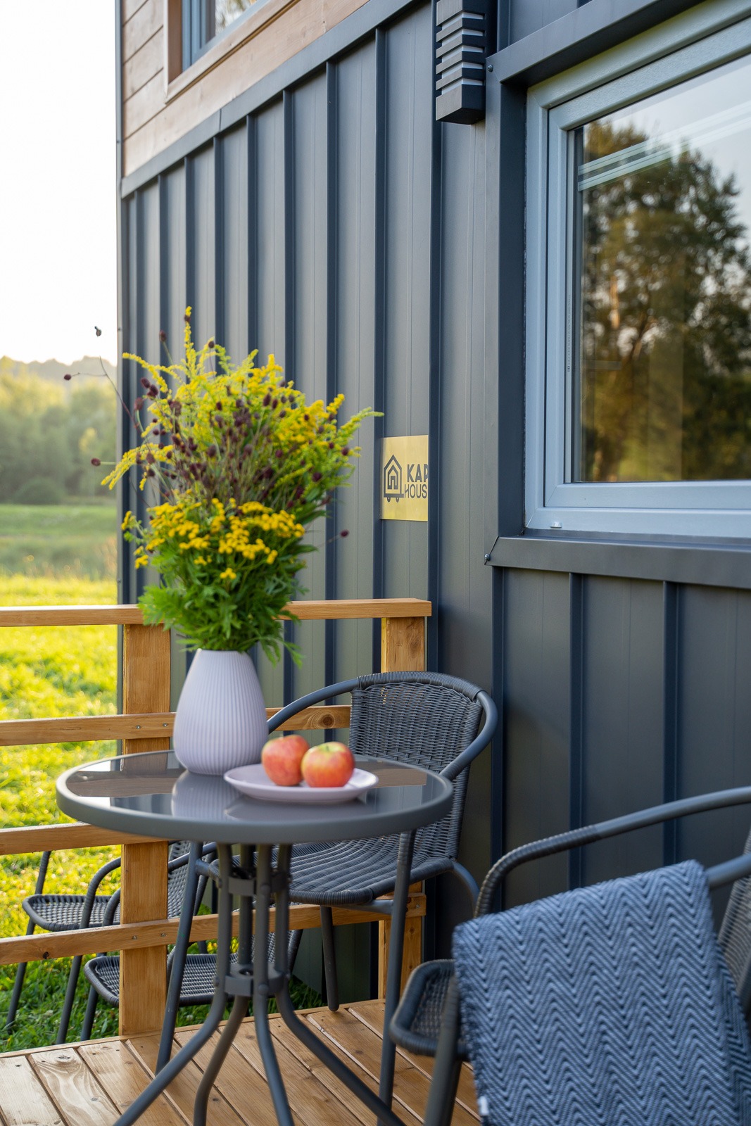 Zewnętrzny taras domu z szarą elewacją z metalu, na drewnianej podłodze stoi okrągły stolik z dwoma jabłkami na talerzu, w wazonie bukiet żółtych kwiatów, w tle widoczne zielone drzewa i pola,...