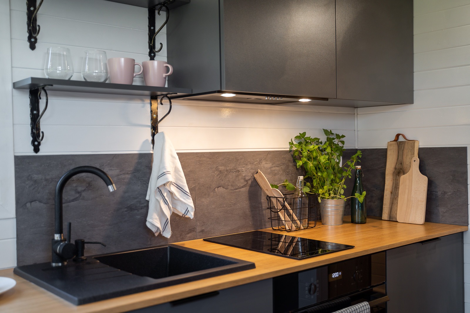 Nowoczesna kuchnia z czarnym zlewem, drewnianym blatem, szarymi szafkami i zielonymi ziołami w metalowej doniczce, oświetlona punktowymi lampami podszafkowymi.