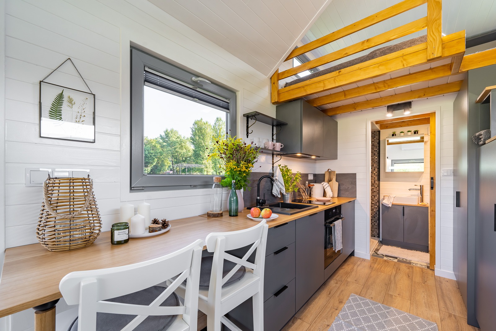 Jasne wnętrze domu z antresolą, widok na kuchnię z szarymi szafkami i drewnianym blatem, białe krzesła barowe, okno z widokiem na zieleń, dekoracje i wejście do łazienki.