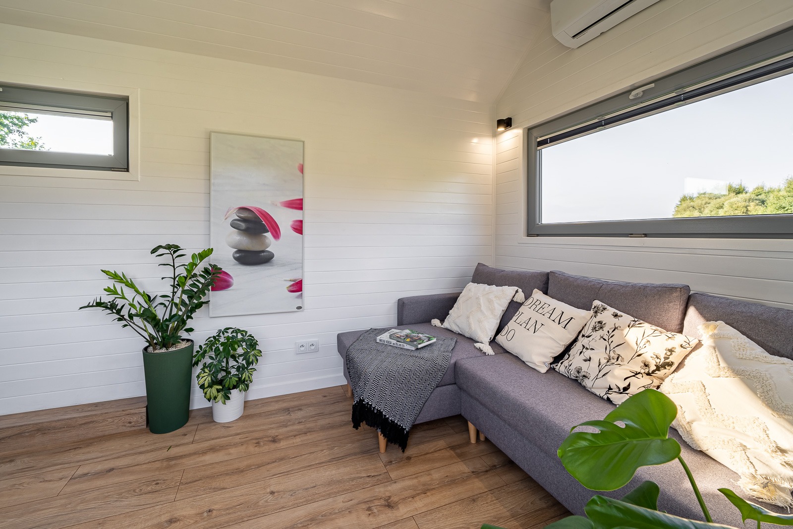 Jasne wnętrze pokoju z drewnianą podłogą, szarym narożnikiem z poduszkami, roślinami w doniczkach i obrazem na ścianie z paneli, klimatyzacja pod sufitem.