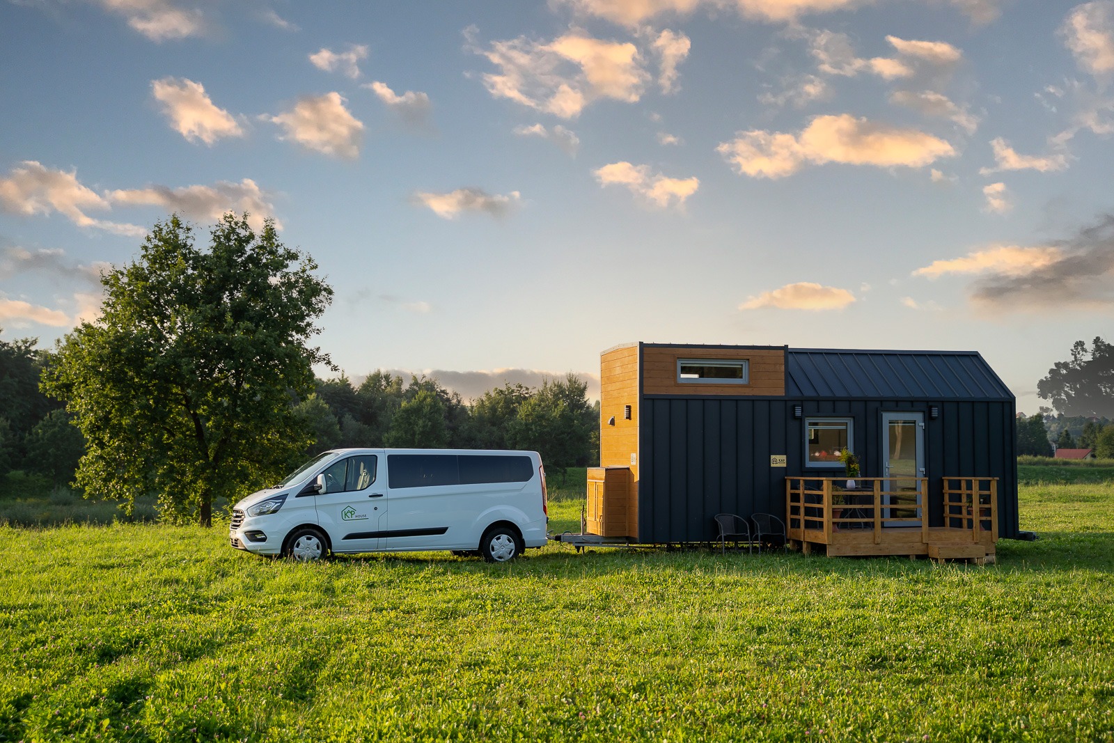 Mobilny, nowoczesny domek na kółkach z ciemną elewacją i drewnianymi akcentami, zaparkowany na zielonej łące obok białego vana z logo firmy.