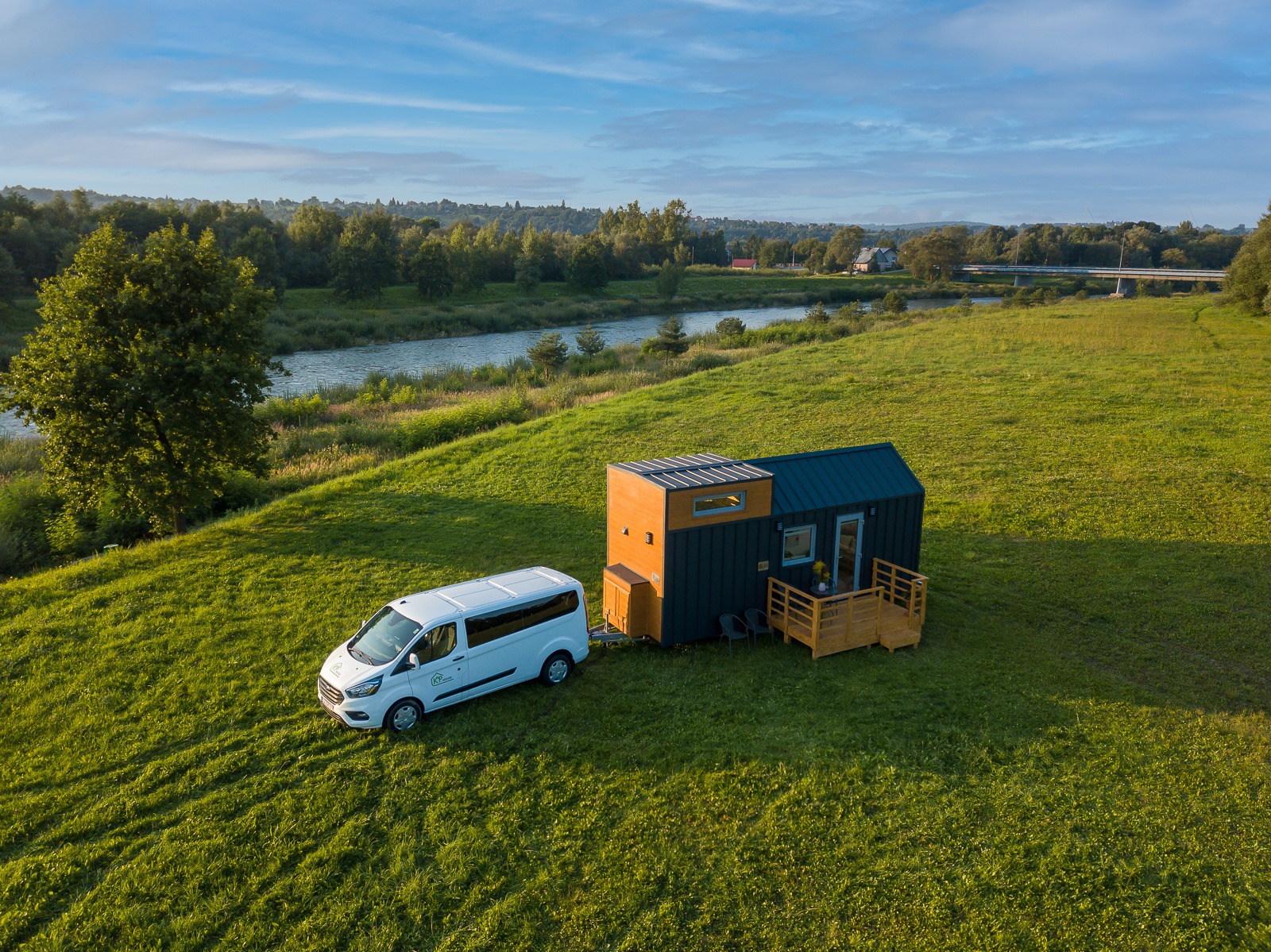 Mobilny, nowoczesny dom szkieletowy na przyczepie, z drewnianym tarasem i białym vanem zaparkowanym obok, na tle zielonej łąki i rzeki.