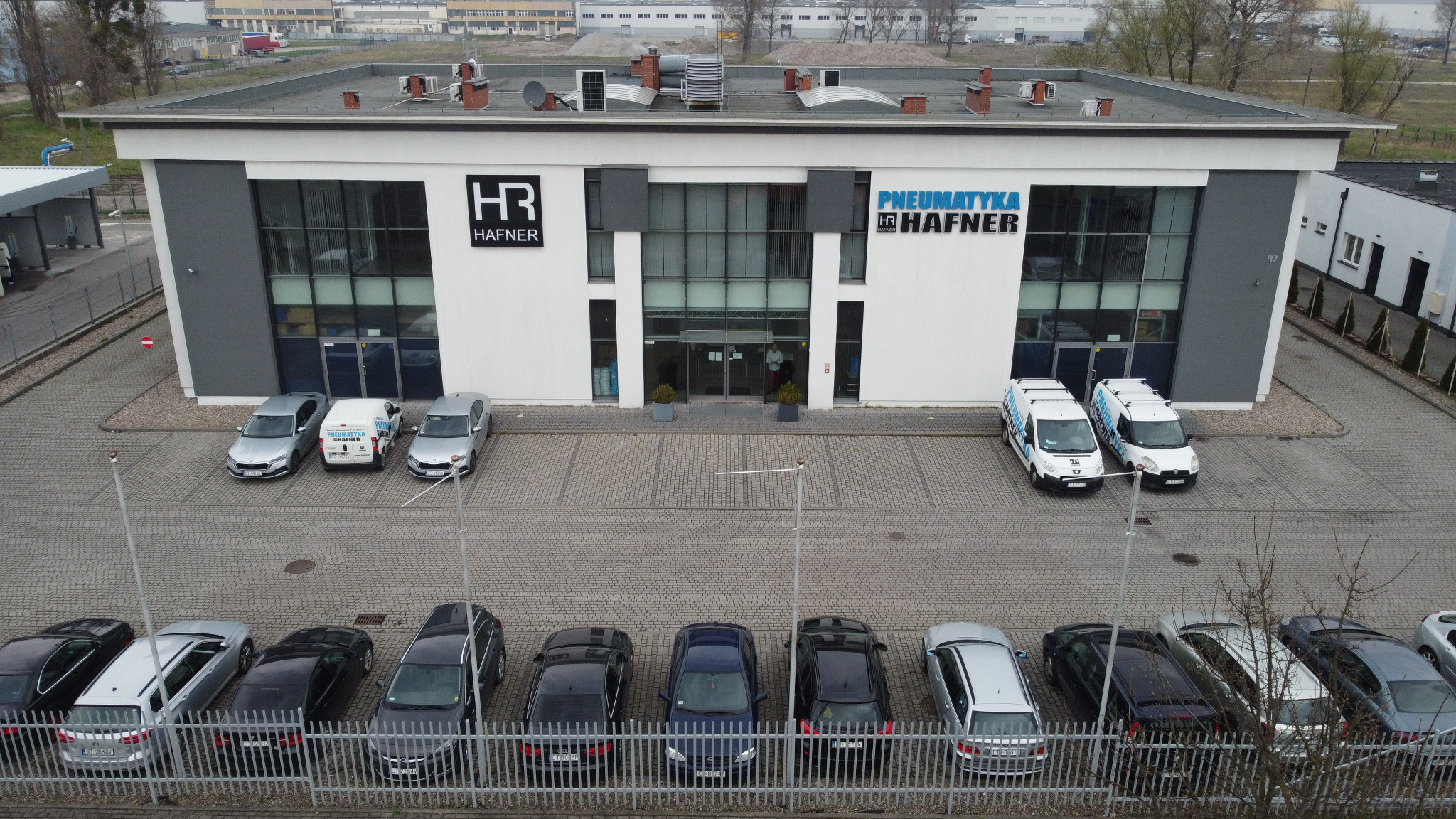 Widok z góry na budynek firmy Hafner z zaparkowanymi samochodami osobowymi i dostawczymi z logo firmy, szary bruk na parkingu, metalowe ogrodzenie na pierwszym planie, pochmurny dzień.