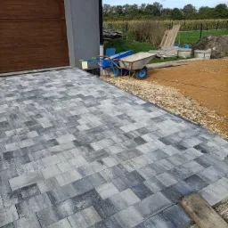 Ułożona szara kostka brukowa przed garażem z brązową bramą, niebieska taczka z zielonymi rączkami stoi obok, widoczne przygotowane podłoże z kamieni i ziemi.