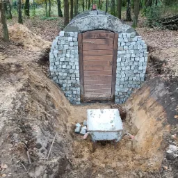 Ziemianka z kamienną fasadą i drewnianymi drzwiami, w trakcie budowy, widoczna taczka z resztkami materiałów, otoczona wykopem i lasem.