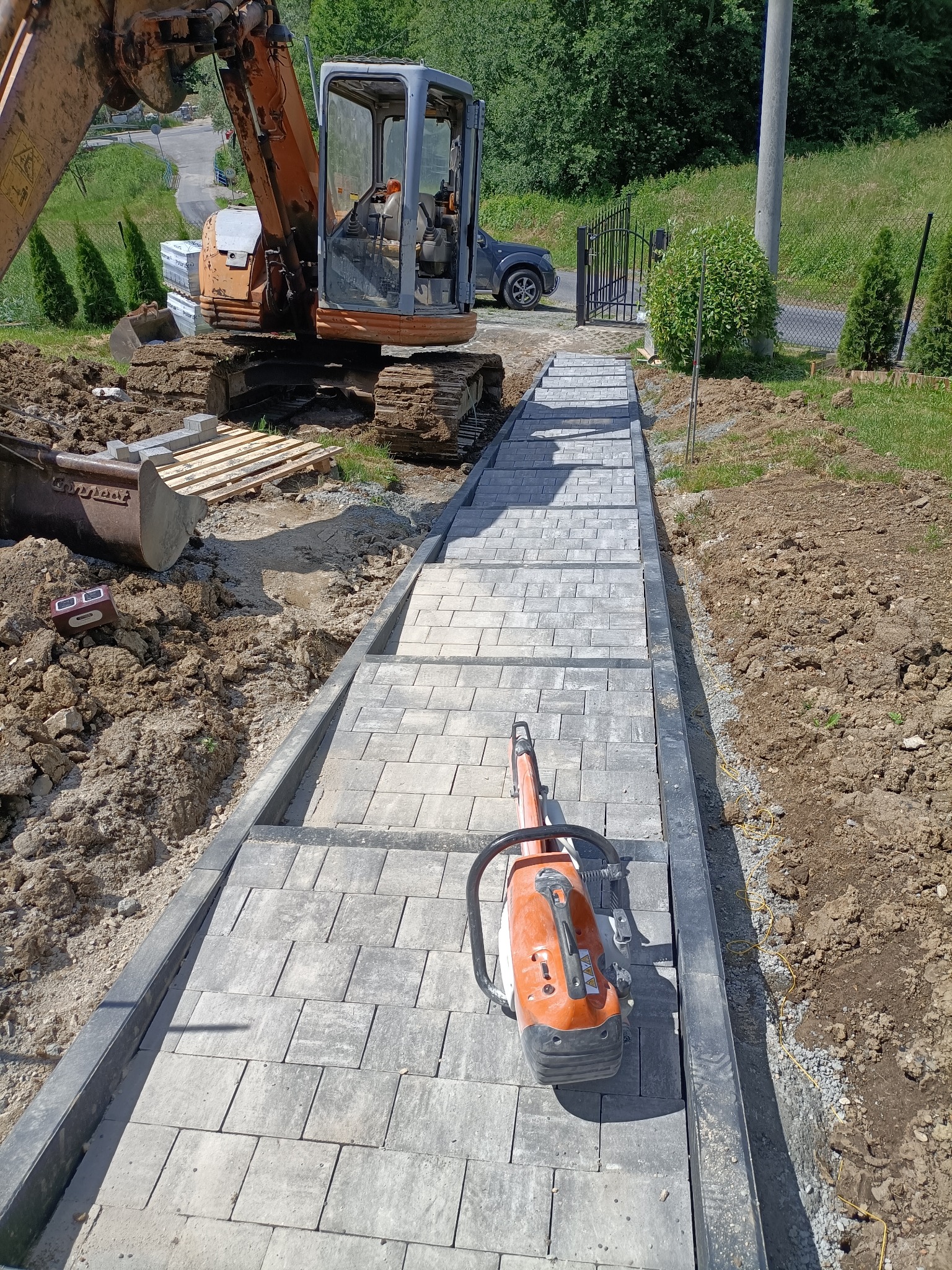 Budowa schodów zewnętrznych z szarej kostki brukowej na podbudowie, z użyciem zagęszczarki wibracyjnej i koparki w tle, widok z góry na częściowo ułożone stopnie.