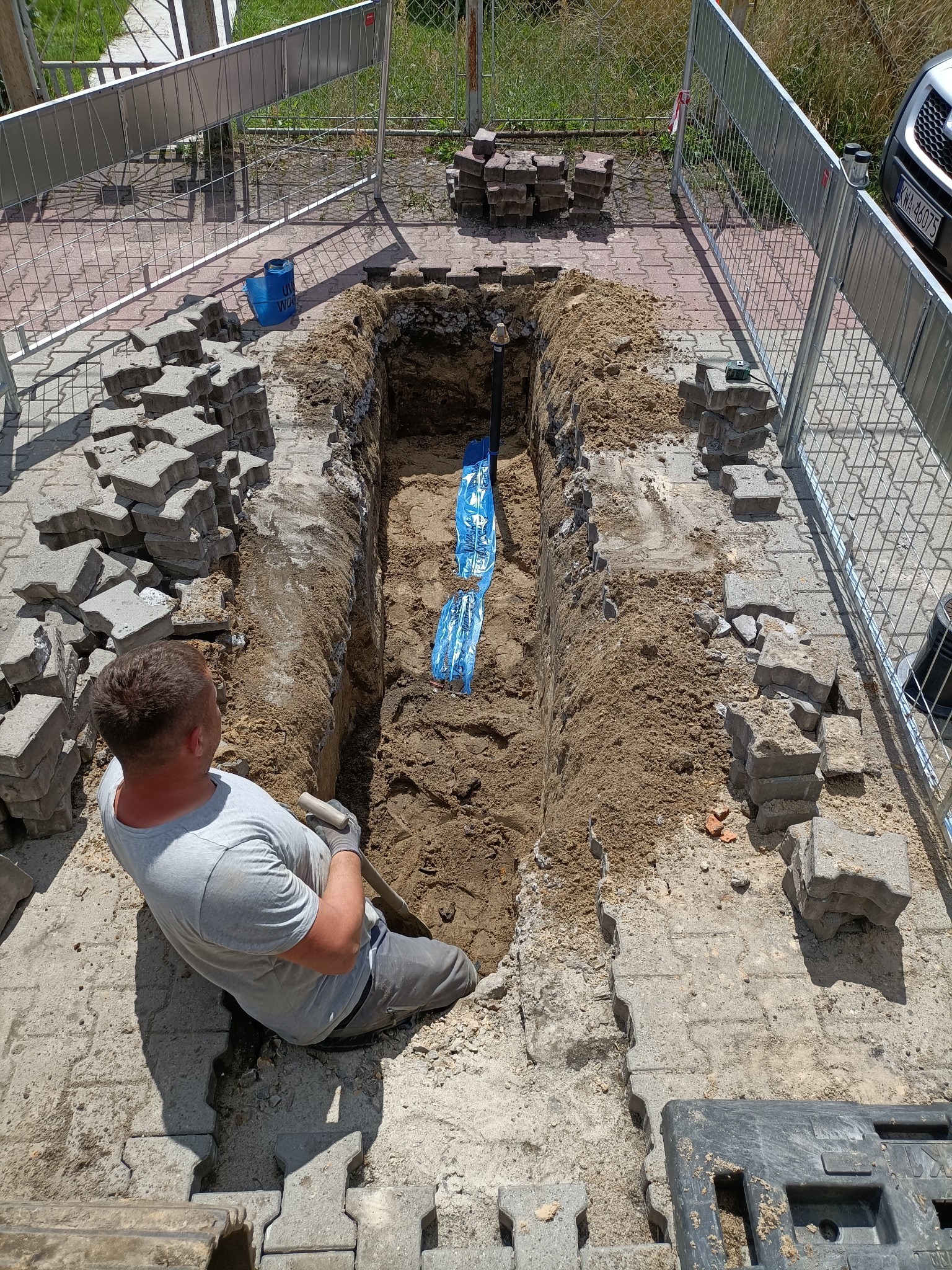 Pracownik siedzi obok wykopu pod rurę na posesji z kostki brukowej, przygotowując się do dalszych prac ziemnych. Widoczny zabezpieczony rów z ułożonymi wzdłuż niego kostkami oraz fragment...