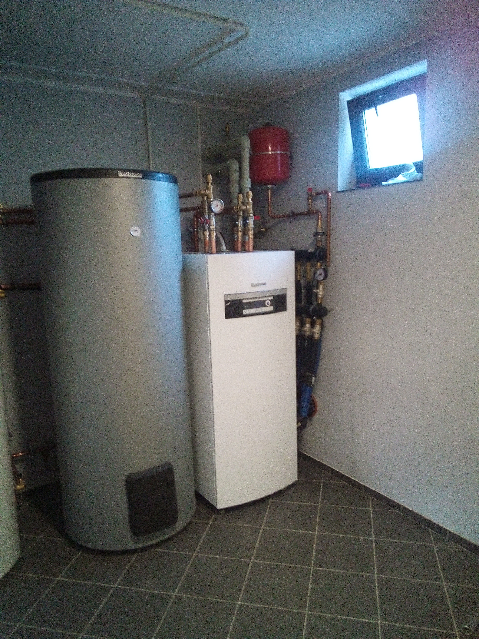 Szary, cylindryczny zasobnik ciepłej wody marki Buderus obok białego kotła grzewczego w kotłowni z ciemnymi, kwadratowymi płytkami podłogowymi i małym oknem.